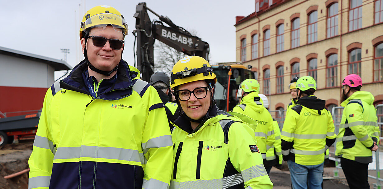 Mattias Sandström och Mariia Zetterberg står intill schaktet med projektgrupppen och grävmaskinen i bakgrunden.